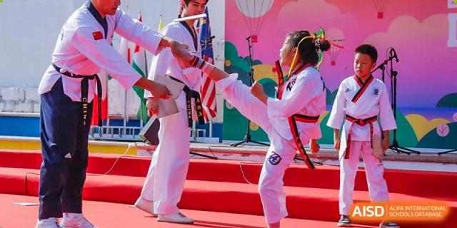A female student in a white Taekwondo uniform practicing a high kick with an instructor during a demonstration at the Qingdao Amerasia International School.