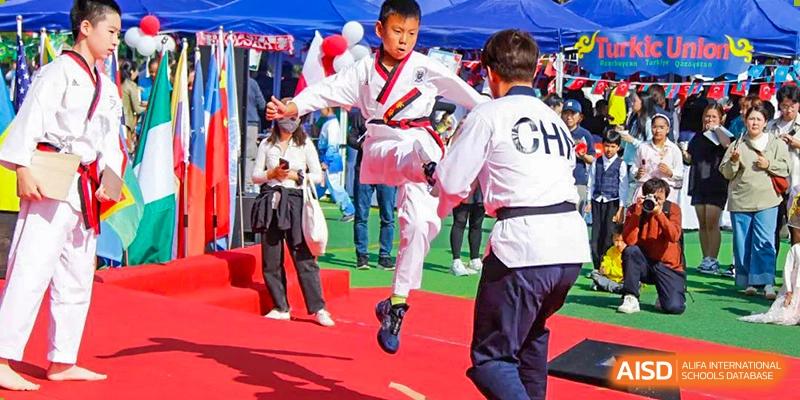 A male student in a white Taekwondo uniform mid-air, performing a flying kick during a demonstration at the Qingdao Amerasia International School.