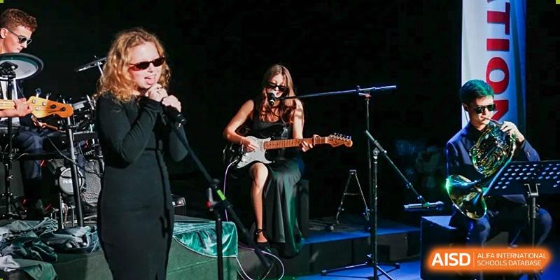 A female musician in a black dress and sunglasses playing an electric guitar on a stage at the Qingdao Amerasia International School.