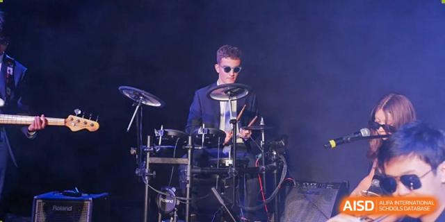 A male musician wearing sunglasses playing the drums on a stage at the Qingdao Amerasia International School.
