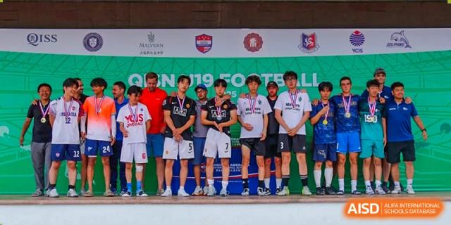 A soccer team posing with their trophies and medals on a podium after a championship at the Qingdao Amerasia International School.