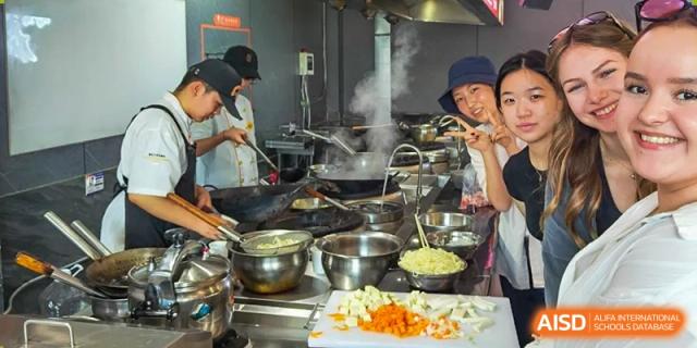 A group of students wearing hats and aprons, cooking in a busy kitchen at the Qingdao Amerasia International School.