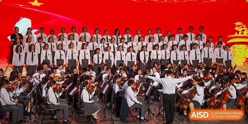 A choir and orchestra from Rugao International Academy BFSU performing on a stage with a red background.