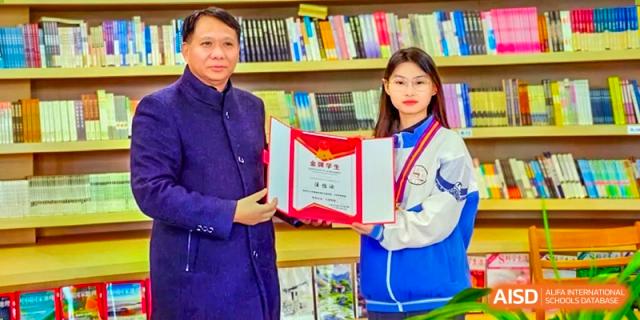 A teacher or staff member presenting a certificate and a red folder to a female student indoors, with a bookshelf visible in the background at Shanghai Wenlai International School.