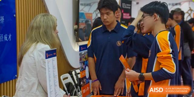 A male student in a dark blue and gold polo shirt stands indoors, holding an orange paper at Shanghai Wenlai International School.