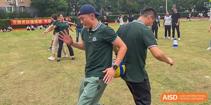 A man in a dark green polo shirt runs while holding a yellow and blue volleyball during an outdoor activity at Shanghai Wenlai International School.