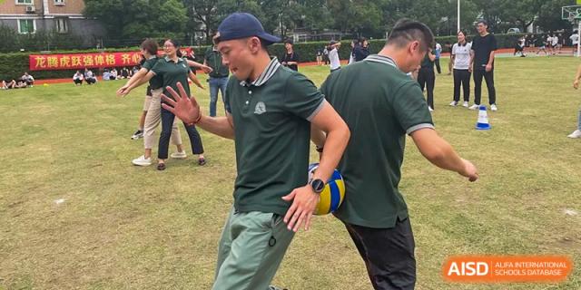 A man in a dark green polo shirt runs while holding a yellow and blue volleyball during an outdoor activity at Shanghai Wenlai International School.