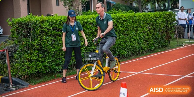 A foreign teacher riding a small yellow bicycle on a track, with a female student walking beside him at an event at Shanghai Wenlai International School.