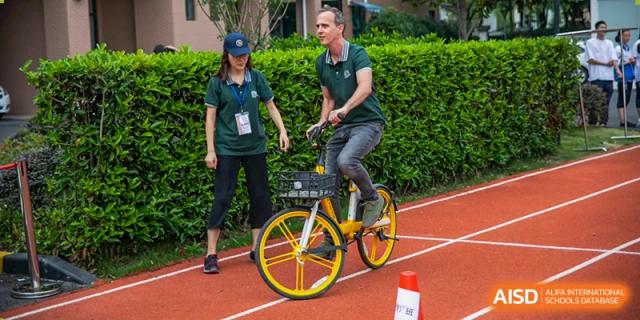 A foreign teacher riding a small yellow bicycle on a track, with a female student walking beside him at an event at Shanghai Wenlai International School.