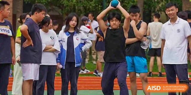 A male student in a black tank top preparing to throw a blue shot put or similar object while other students watch on a red track at Shanghai Wenlai International School.
