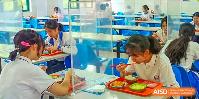 A student eating a meal in a cafeteria while sitting behind a clear plastic sneeze guard for safety at Shanghai Wenlai International School.