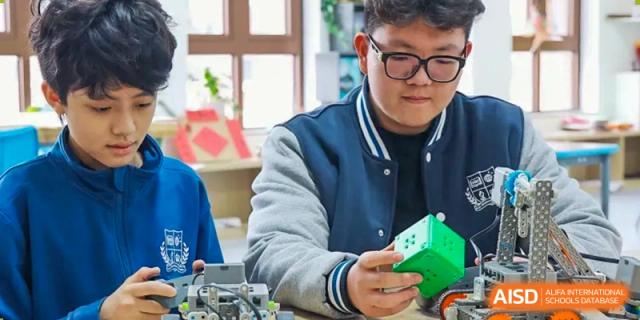 A male student at Sias International School wearing a navy and white jacket works with a small green electronic device at his desk.