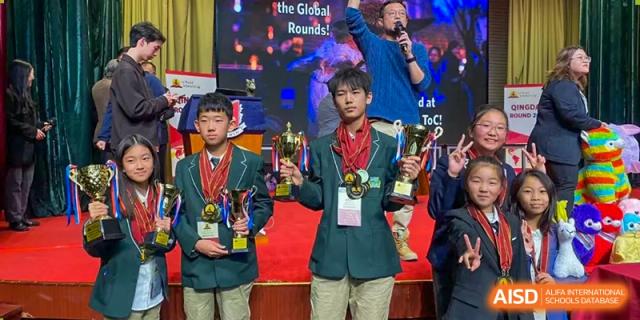 Two students from Sias International School wearing green jackets and medals, holding large trophies on stage at a competition with a crowd and speaker visible in the background.