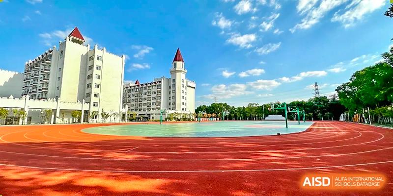 SUZHOU INTERNATIONAL FOREIGN LANGUAGE SCHOOL campus with a white building that has a red conical roof next to a red track and a multicolored sports court under a blue sky with clouds.