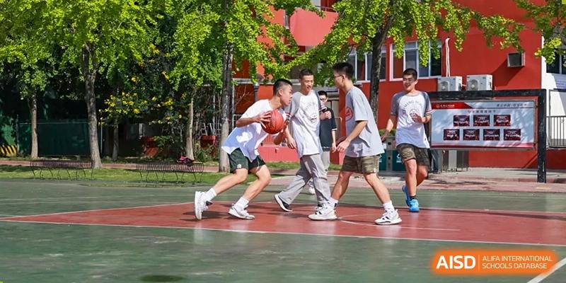 Two Weiming School Chengdu Qingbaijiang students dribble a basketball in a one-on-one game on an outdoor court surrounded by trees.