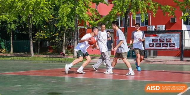 Two Weiming School Chengdu Qingbaijiang students dribble a basketball in a one-on-one game on an outdoor court surrounded by trees.