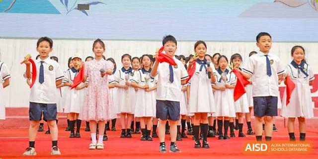 Young Weiming School Chengdu Qingbaijiang students in school uniforms stand on a stage, with one student in front wearing a red scarf and holding a flag.