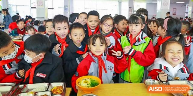 A group of smiling students from Weiming School Chengdu Qingbaijiang posing with their food in the cafeteria.