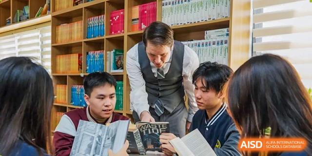 A male teacher in a vest and tie leaning over two male students, pointing to text in a book in a library or classroom at Weiming School Shenzhen Nanshan.