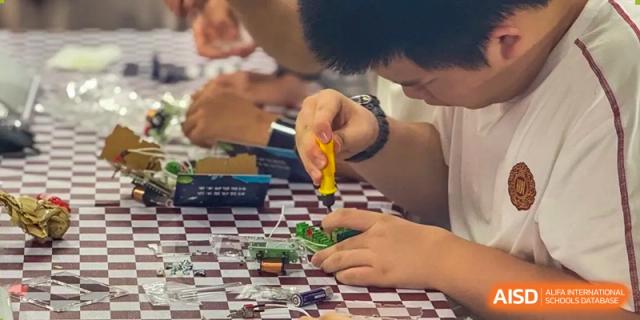 A close-up shot of a student wearing glasses, carefully working with a soldering iron and electrical components on a circuit board at Weiming School Shenzhen Nanshan.