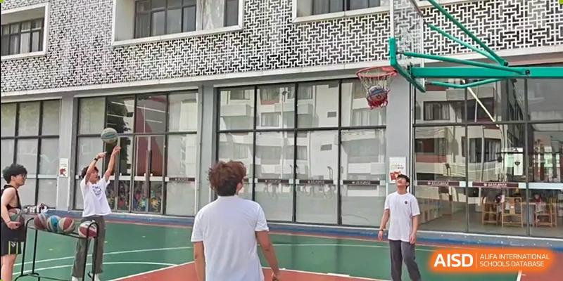 A student in a white shirt shooting a basketball at an outdoor hoop near a modern building at Weiming School Shenzhen Nanshan.