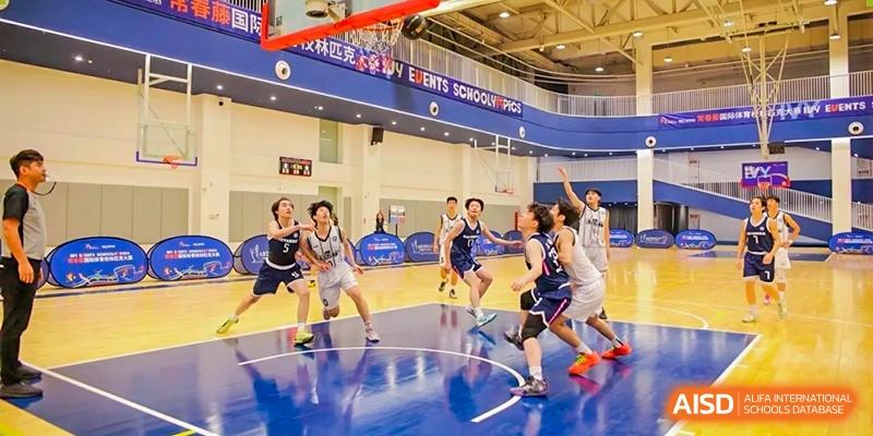 Students playing an intense game of basketball on an indoor court, with spectators visible in the background, at Weiming School Shenzhen Nanshan.