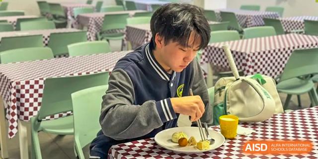 A student in a blue and black jacket eating a small portion of food from a plate in the school dining area at Weiming School Shenzhen Nanshan.