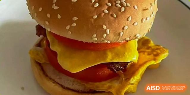 A close-up of a large, fresh cheeseburger with a sesame bun, tomato, and lettuce, served at Weiming School Shenzhen Nanshan.