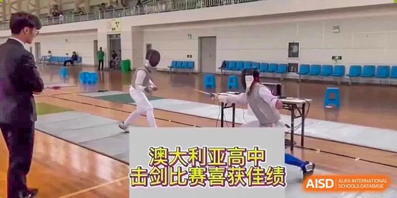 Two students fencing in a gymnasium with a large screen displaying Chinese text, showing a sports competition at Wenzhou No.21 Middle School.