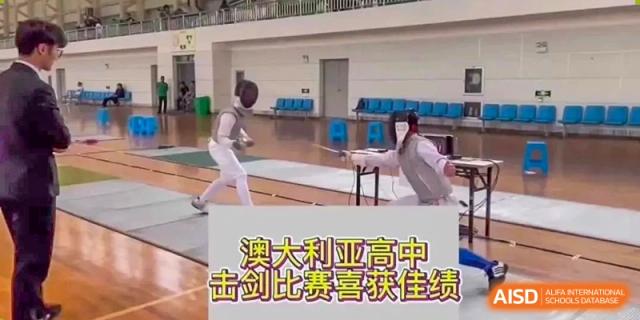 Two students fencing in a gymnasium with a large screen displaying Chinese text, showing a sports competition at Wenzhou No.21 Middle School.