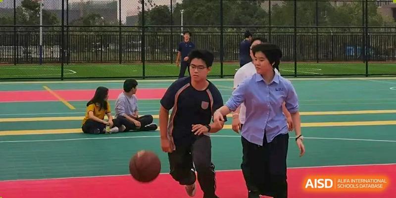 Two male students playing basketball outdoors on a court during a physical education class or break time at Wuhan Optics Valley ULINK School.