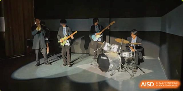 Students performing on electric guitars and drums in a dark practice room or band studio at Wuhan Optics Valley ULINK School.