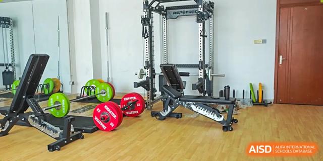 A well-equipped indoor weight training and fitness area with a squat rack, bench, and various colored weight plates on a wooden floor at Wuhan Optics Valley ULINK School.
