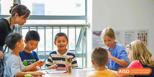 A smiling Wuhan Yangtze International School elementary student working on an assignment with a pencil at a table in a bright classroom.