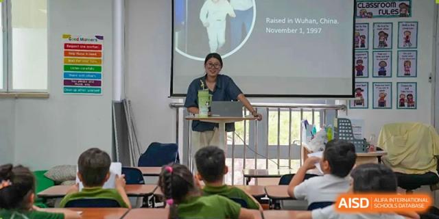 A Wuhan Yangtze International School teacher standing at a podium and lecturing to students seated on the floor and at tables during a presentation.