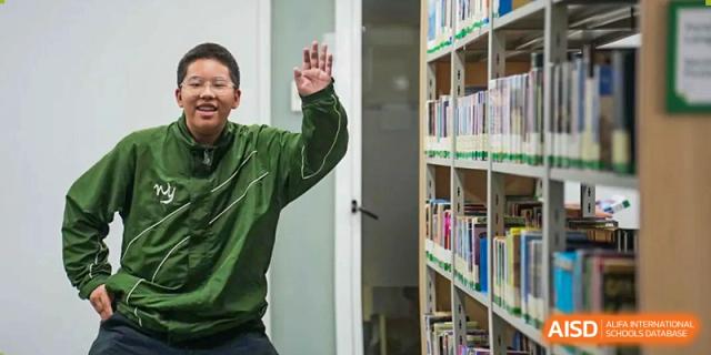 A Wuhan Yangtze International School student in a green sweatshirt raising a fist in excitement in a hallway next to bookshelves.