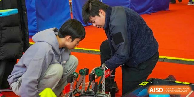 Two Wuhan Yangtze International School students collaborate closely on adjusting a mechanism on a black and silver robotics project.