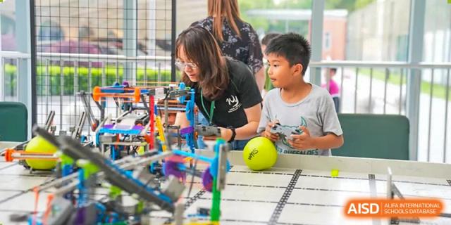 A Wuhan Yangtze International School teacher smiles while guiding two students working with colorful LEGO robotics components on a table.