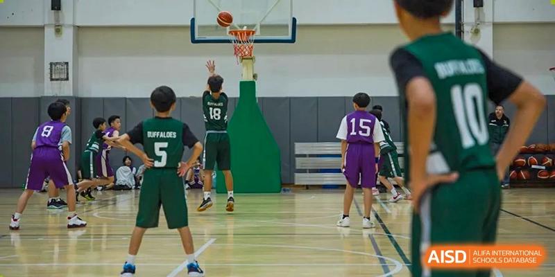 Two Wuhan Yangtze International School students playing basketball in a gym, with one student in a green jersey shooting a layup and another in a purple jersey looking on.
