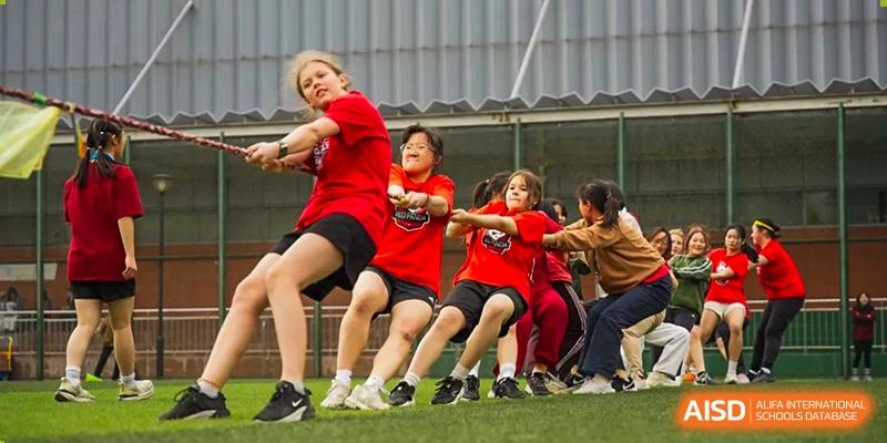 A team of Wuhan Yangtze International School students in red shirts enthusiastically pulling a rope during a tug-of-war competition on a field.