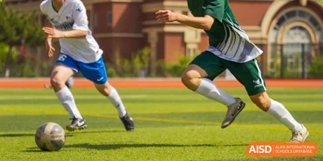 A close-up of a Wuhan Yangtze International School athlete's lower body kicking a soccer ball on a green turf field.