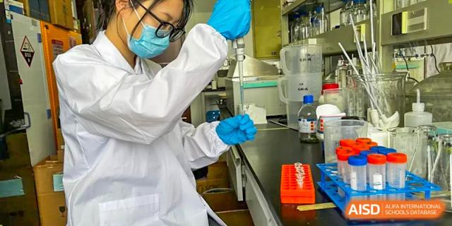 Two students in lab coats, gloves, and safety masks working with equipment and chemicals in a science laboratory at Wuxi Dipont School of Arts and Science.