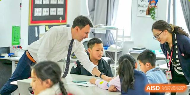 A male teacher in a tie helping two young students with a hands-on activity or assignment in a brightly lit classroom at Wuxi Dipont School of Arts and Science.
