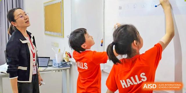 Two young students in orange shirts writing or drawing on a vertical whiteboard in a classroom at Wuxi Dipont School of Arts and Science.
