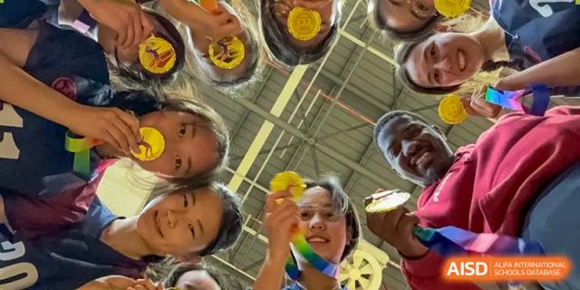 A group of students looking up and smiling, with one holding a yellow ball, in an indoor sports facility at Wuxi Dipont School of Arts and Science.