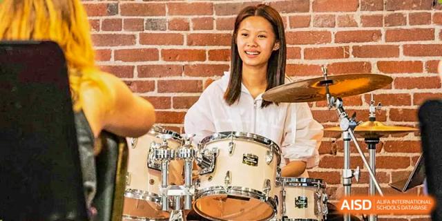 A smiling female student in a white shirt sitting behind a drum set and playing at Wuxi Dipont School of Arts and Science.