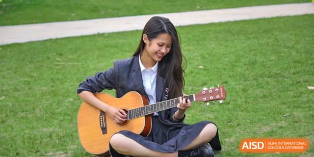 A female student in a school uniform sitting on the grass outdoors and playing an acoustic guitar at Wuxi Dipont School of Arts and Science.