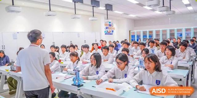 A large classroom of students in white lab coats seated at desks, possibly conducting an experiment or attending a science lecture, at Wuxi Kuangyuan Bilingual School.