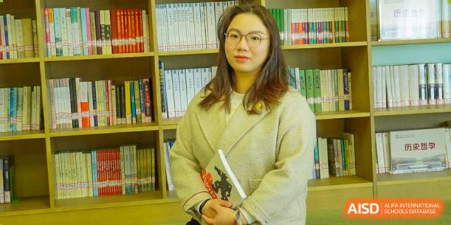 A female student or teacher wearing glasses and a light coat, holding a book and looking directly at the camera in a library setting at Wuxi Kuangyuan Bilingual School.