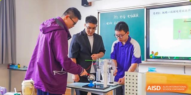 A teacher in a suit working with two male students on a physics or science experiment using lab equipment at Wuxi Kuangyuan Bilingual School.